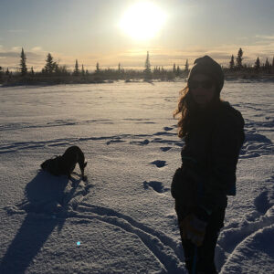 Kim Dahm stands in silhouette while her dog plays in the snow at sunset
