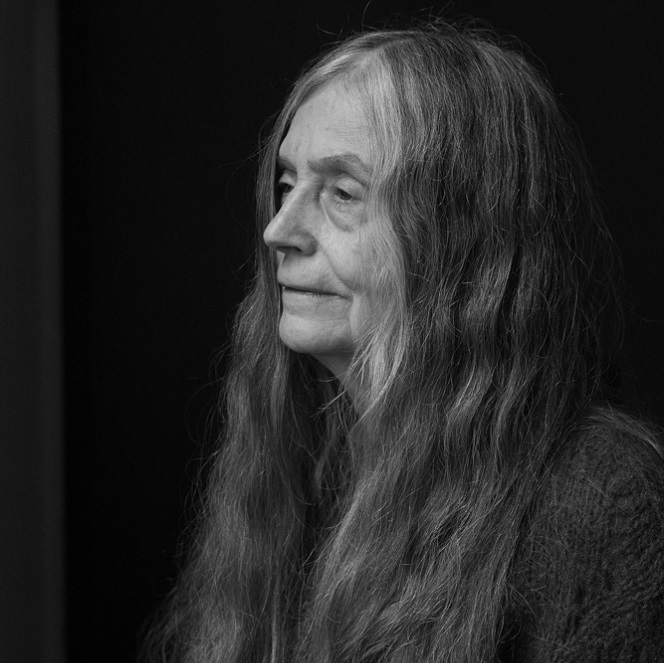 A black and white photograph of a white woman with long, gray hair, looking off to the left, against a black background