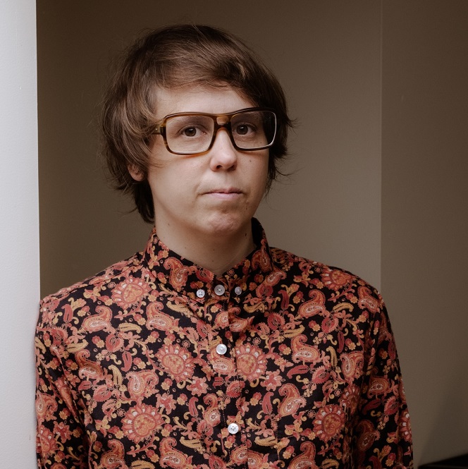 A person with short blond hair and large glasses, wearing a mauve paisley shirt, looks into the camers