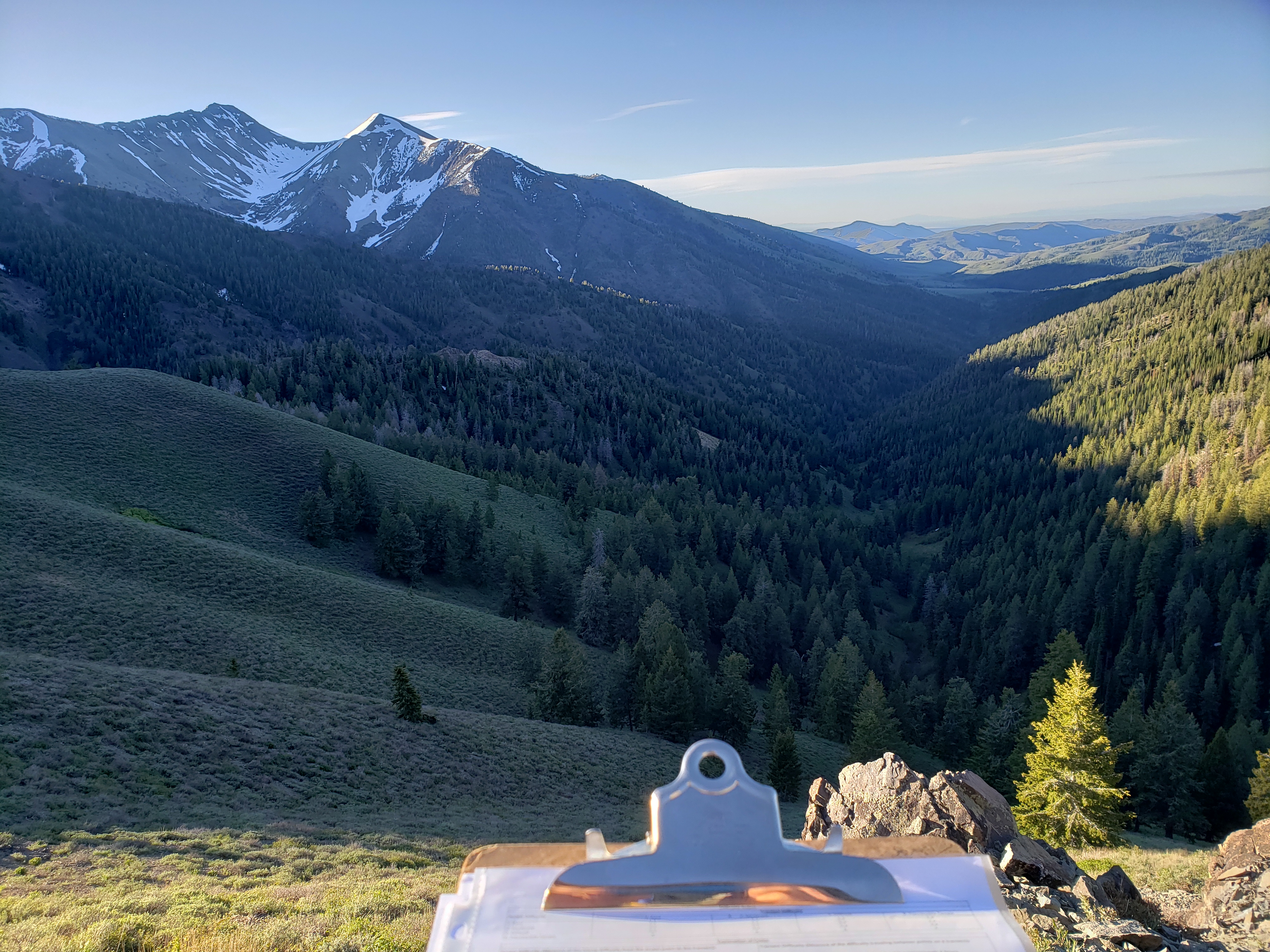 part of a clipboard and data sheet are visible in the foreground. Sagebrush hills, forest, and snowy mountains show in the background