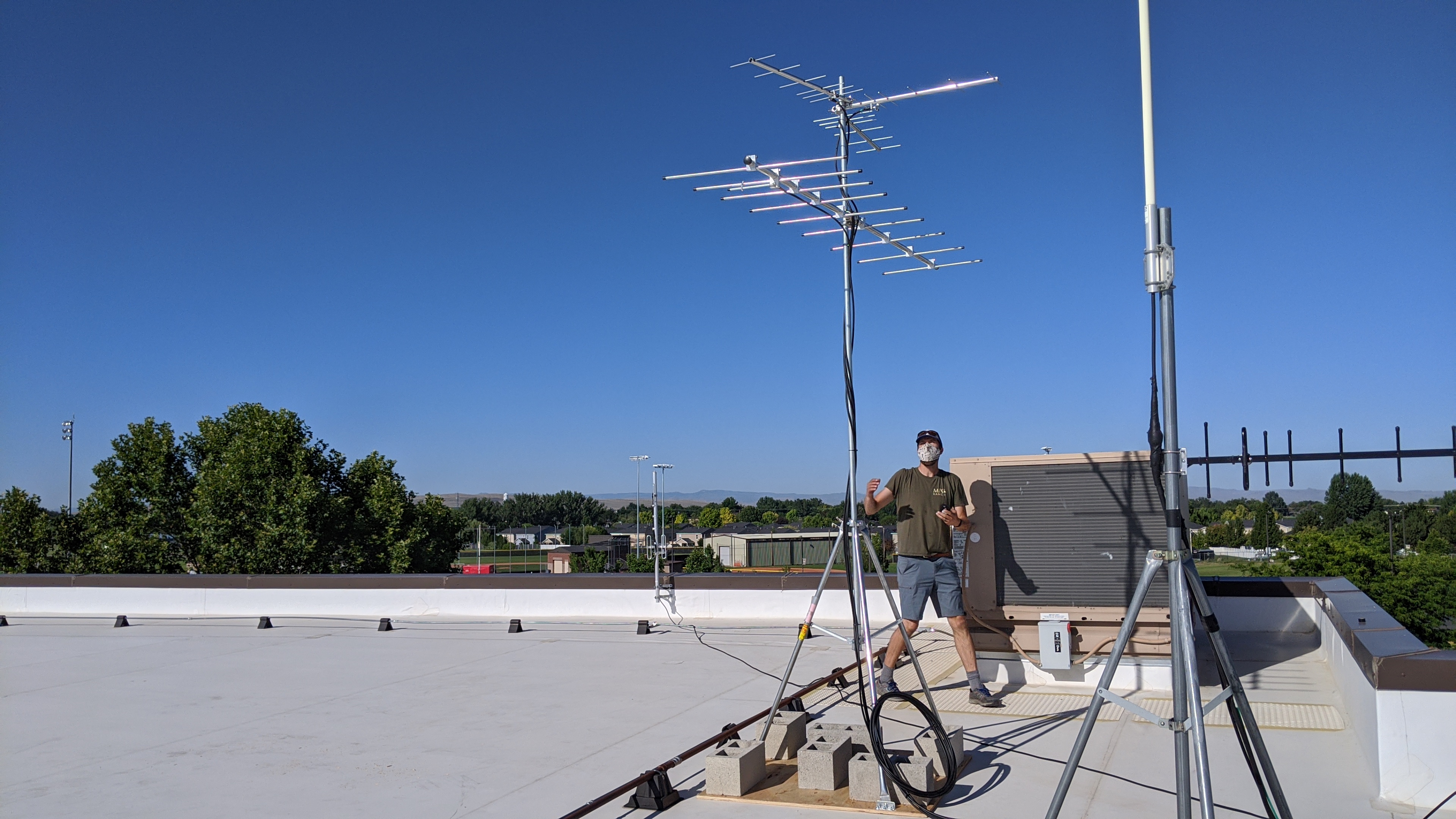 a biologist wearing a cloth face mask stands on the flat roof of a building looking up at an antenna array and unfurling a wire attached to the antenna
