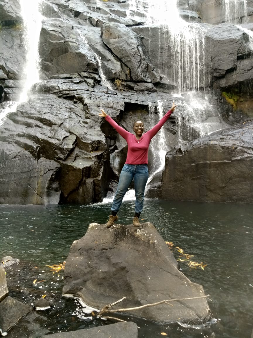 A black woman stands with arms raised joyfully in front of a cascading waterfall