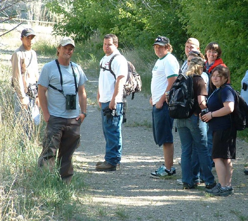 two smiling biologists stand outdoors with a group of college age students
