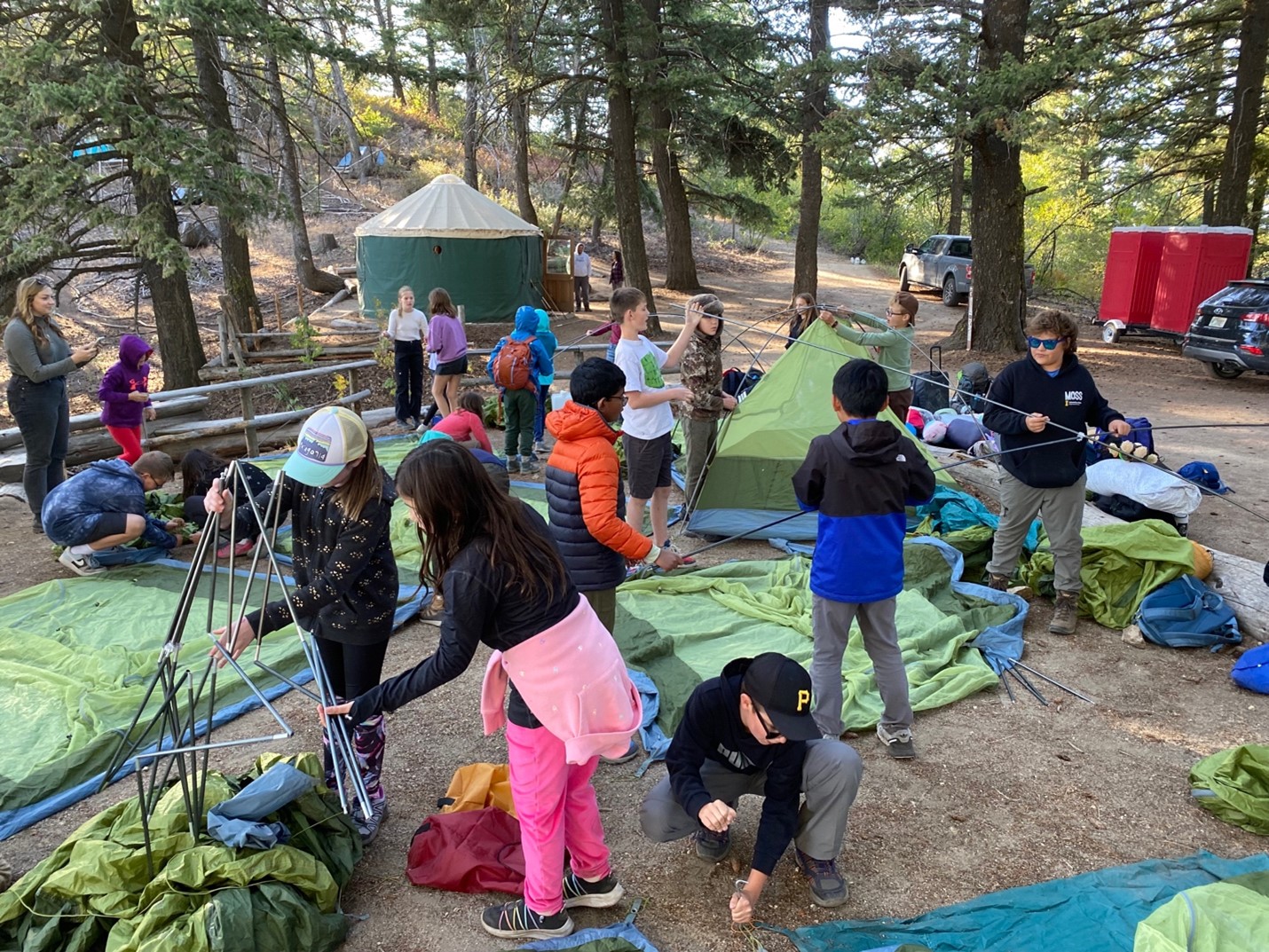 a chaotic scene of twenty fifth graders packed together on the main camping pad at Lucky Peak. each working on a different camp setup task. Some are working together on detangling tent poles, some are pounding tent stakes, some are laying out tents. The Lucky Peak yurt, douglas fir trees, and subarus are in the background