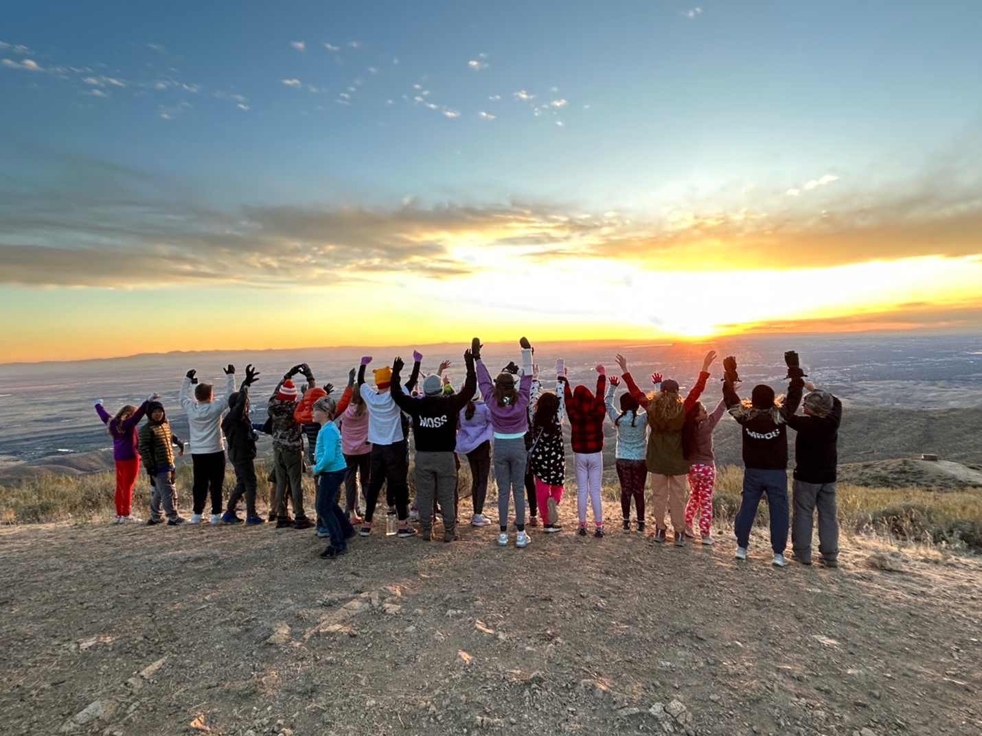 a group of about 20 students and a few parents stand at the top of Lucky Peak overlooking Boise. They are facing away over the valley, raising their arms into the air in celebration. The sun is setting with a beautiful orange glow
