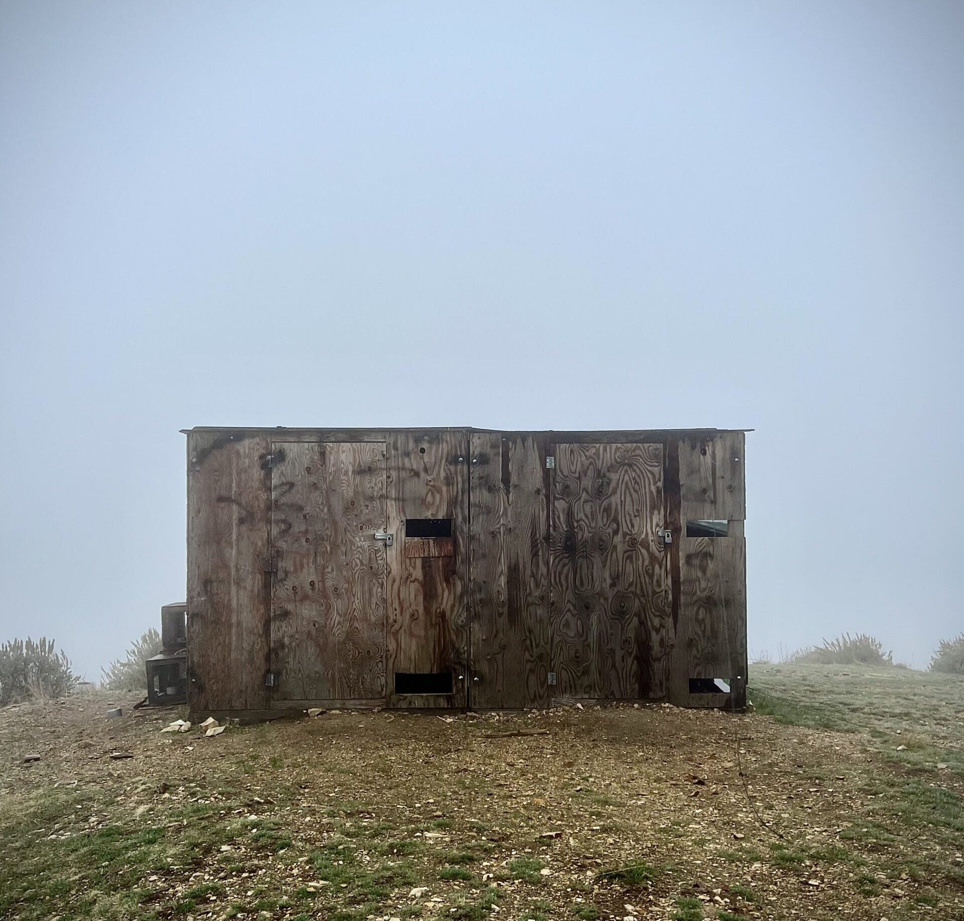 in the foreground is the wooden trapping blind (like a small plywood box) sitting on a gravely hilltop. The surrounding background is completely socked in with fog, with no terrain or features visible