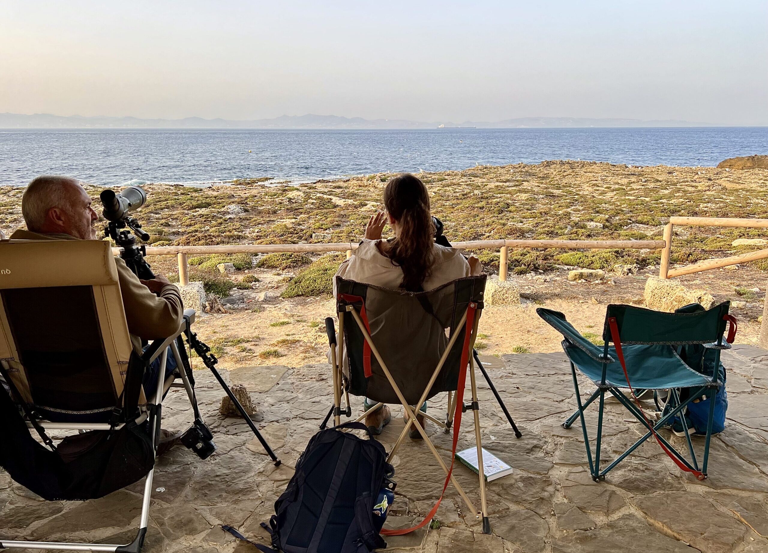 Two people seen from the back sit in foldable camping chairs. They are looking out across a rocky beach and water, with distant mountains barely seen in the haze across the strait. They have spotting scopes set up on tripods looking out over the water.