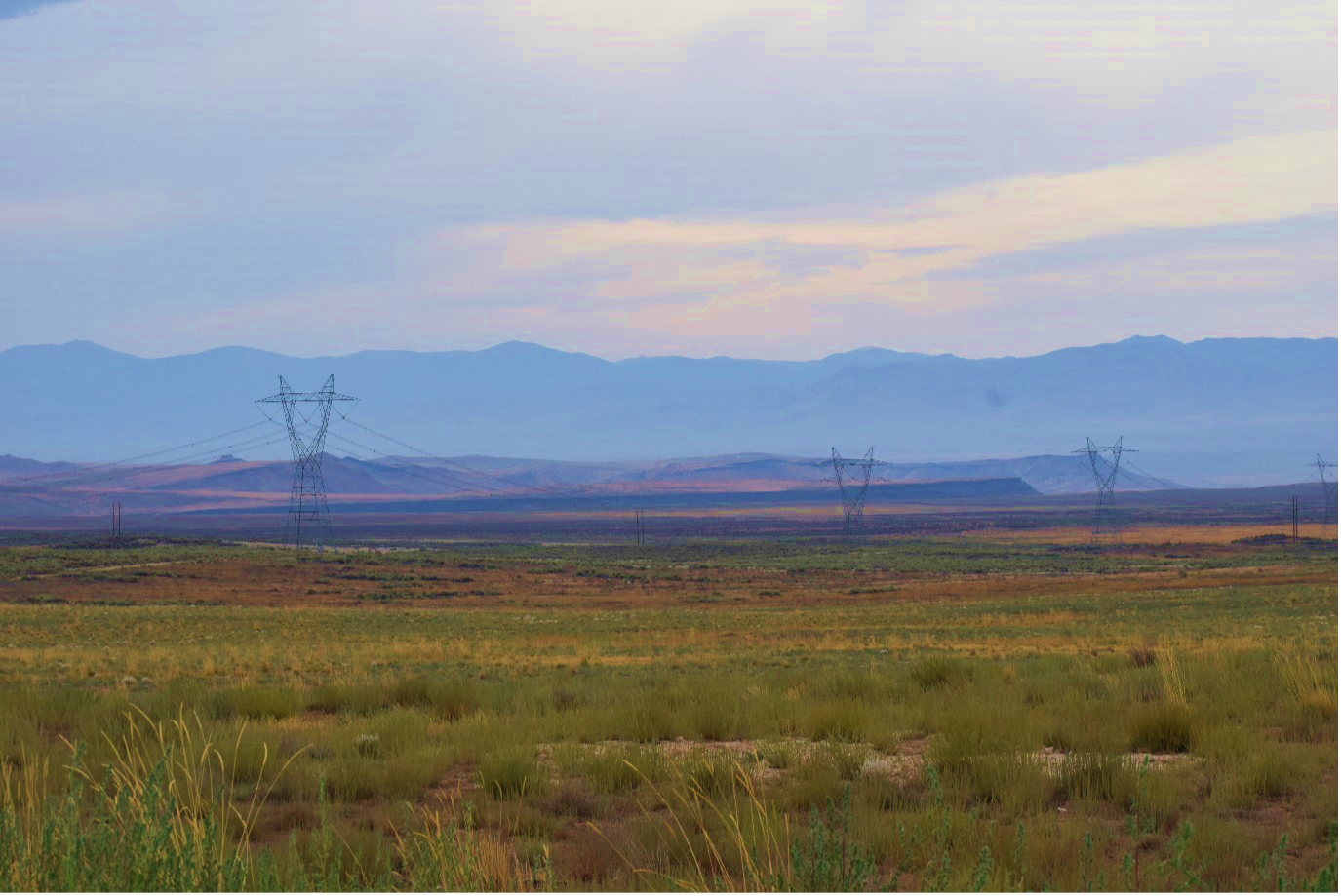 a landscape of mixed grasses and weeds stretches toward mountains and bluffs in the distance. Immense power transmission towers dot the landscape, connected by power lines.