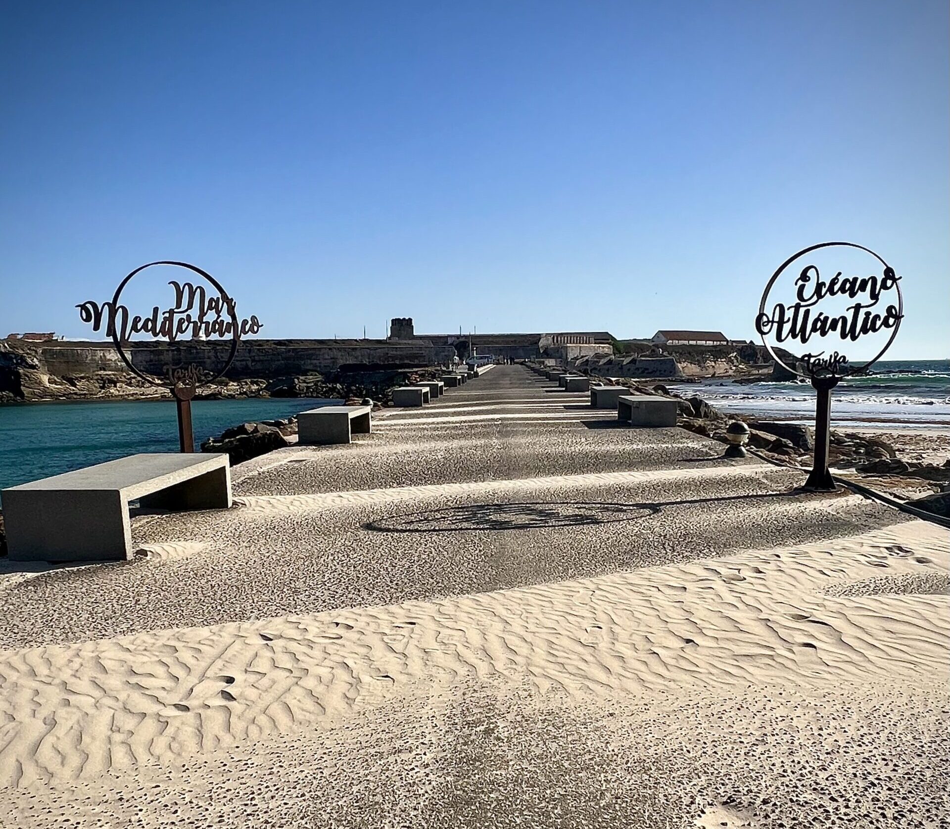A long bridge jetty stretches out away from the camera into the distance, with water on either side. Two signs are on either side, with cursive script cut out of metal in a silhouette design. The left sign reads "mar mediterraneo" and the right reads "oceano atlantico"