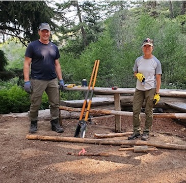 scott and liz stand next to partially built railings. a toolbox, post hole digger, work gloves, and thermos are scattered around the work area