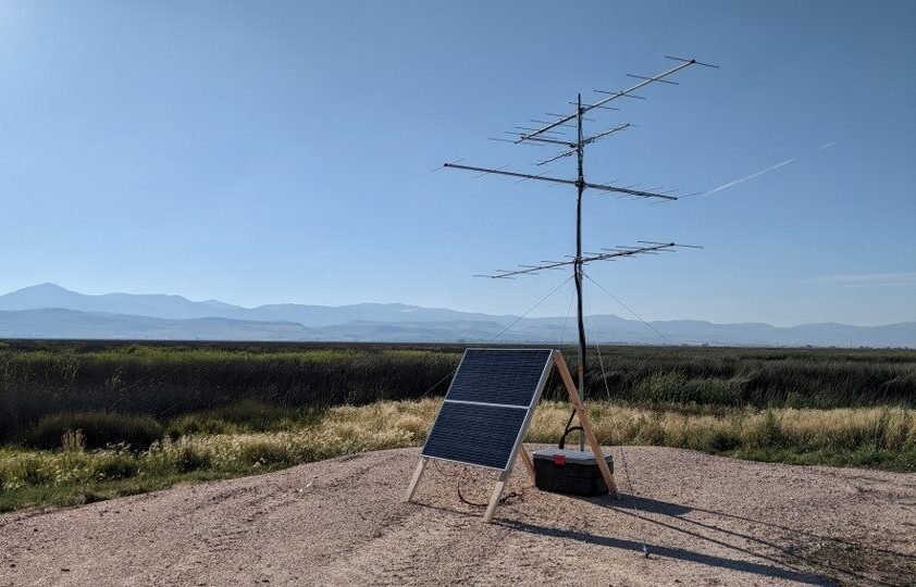 a view of a Motus antenna and solar panel setup. The antenna looks like the old TV antennas people used to have at their houses. A somewhat T shape with some smaller crossbars. The solar panel sits on an A frame at the base of the antenna. In the distance you can see Bear Lake refuge with grasses and marshy cattails, with mountains in the hazy distance