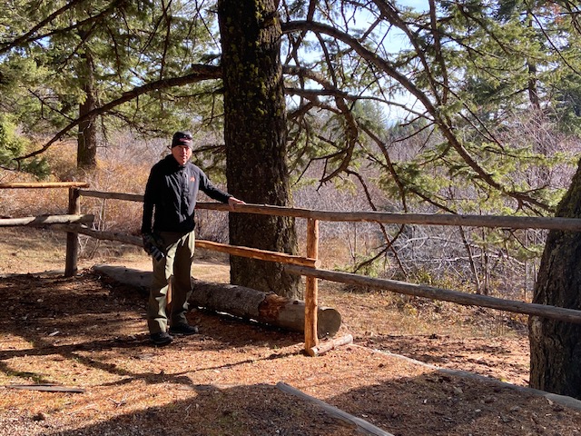 Clyde stands next to a railing built from raw douglas fir logs. in the background is Lucky Peak's main camp with douglas fir trees and bitter cherry shrubs. most of the shrubs are leafless in the late fall weather