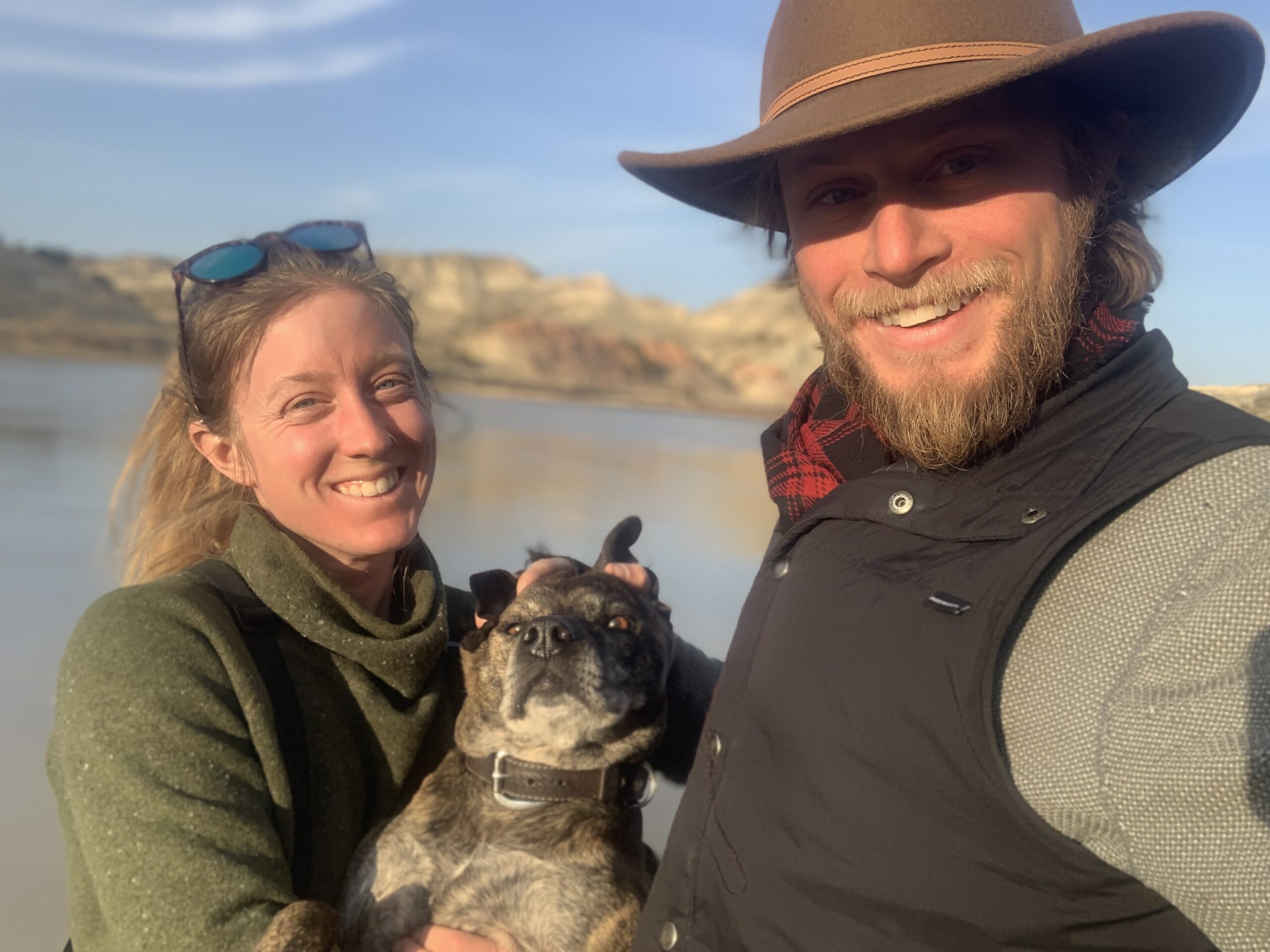 Lesley and Ryan smile for a selfie with their dog. the background reveals sandy cliffs at the shore of a large lake or reservoir