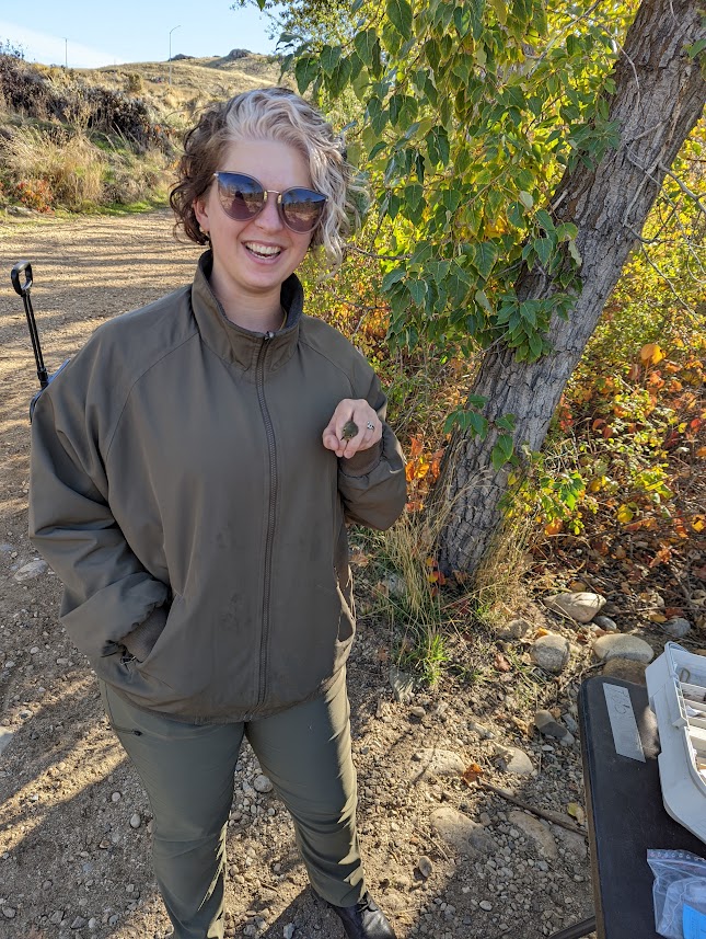 mariah stands smiling and wearing sunglasses near the banding table at the Diane Moore Nature Center. She's wearing olive-green pants, an olive-green jacket, and dark boots. In her hand is a tiny female ruby-crowned kinglet held in banders grip. her head feathers are the same color as Mariah's clothes!