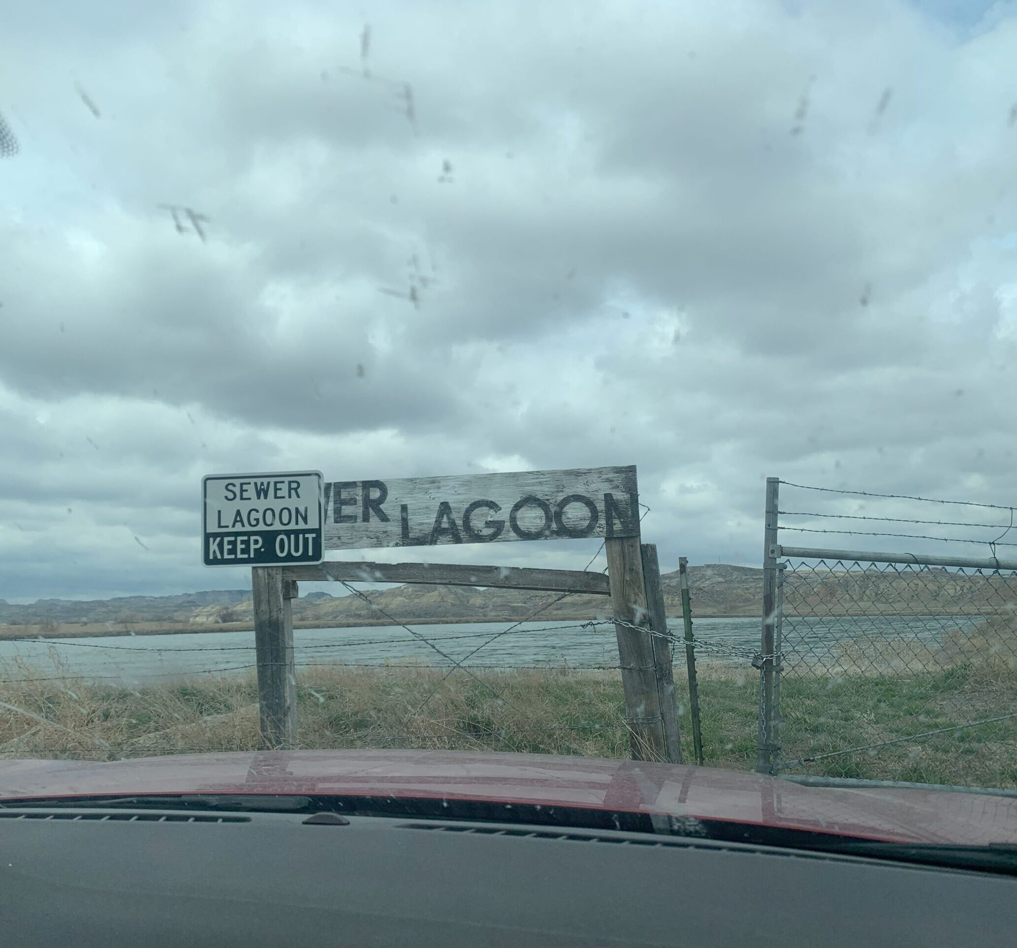 A photo taken through the windshield of a vehicle. There is a locked gate in front of some water that is a sewer lagoon. There are 2 signs stating that it is a sewer lagoon and one says KEEP OUT. The cloudy skies look ominous.