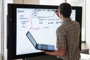 Student holding a laptop and writing on a digital white board.