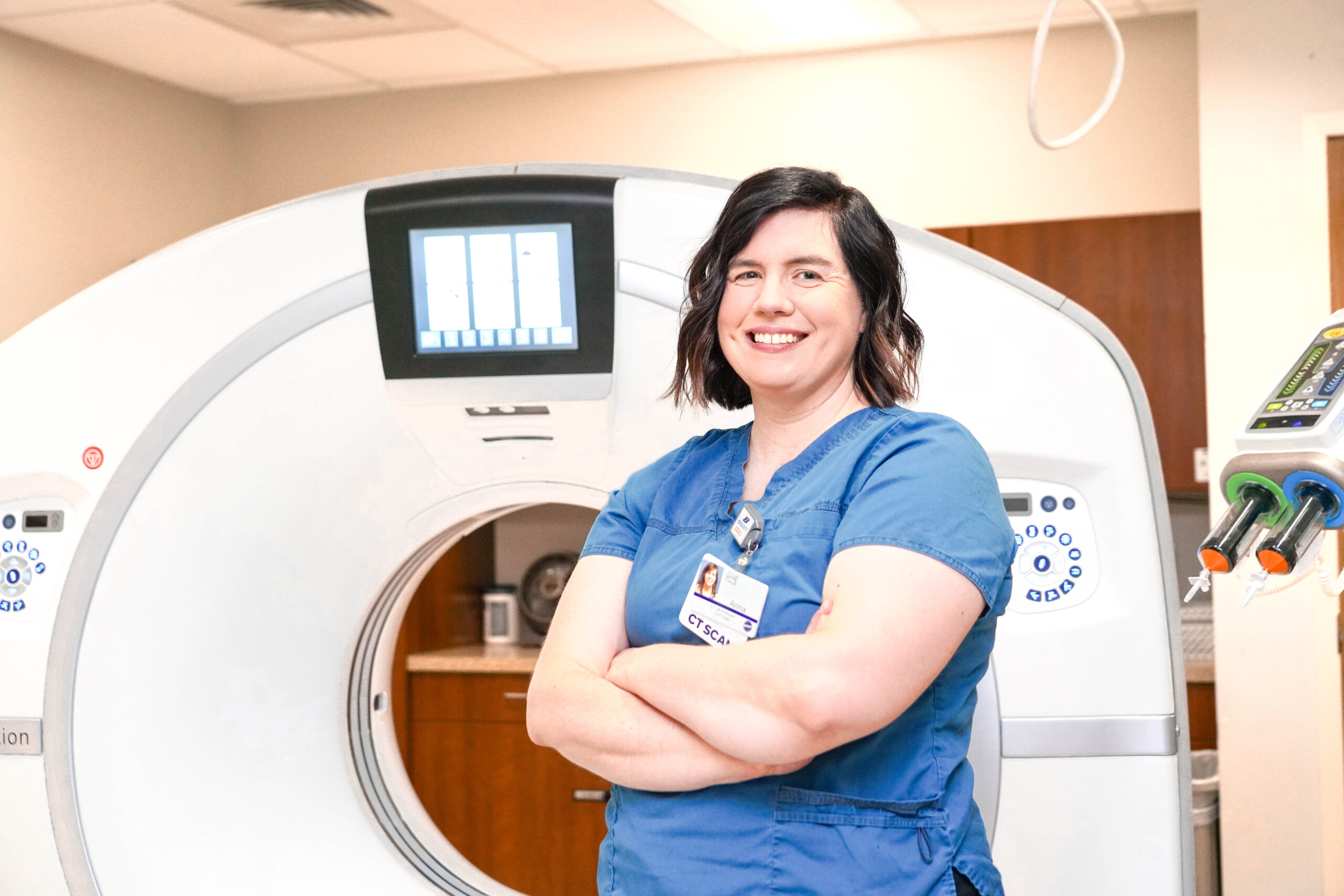 Anna Schreiber wears blue scrubs and stands in front of imaging machine.
