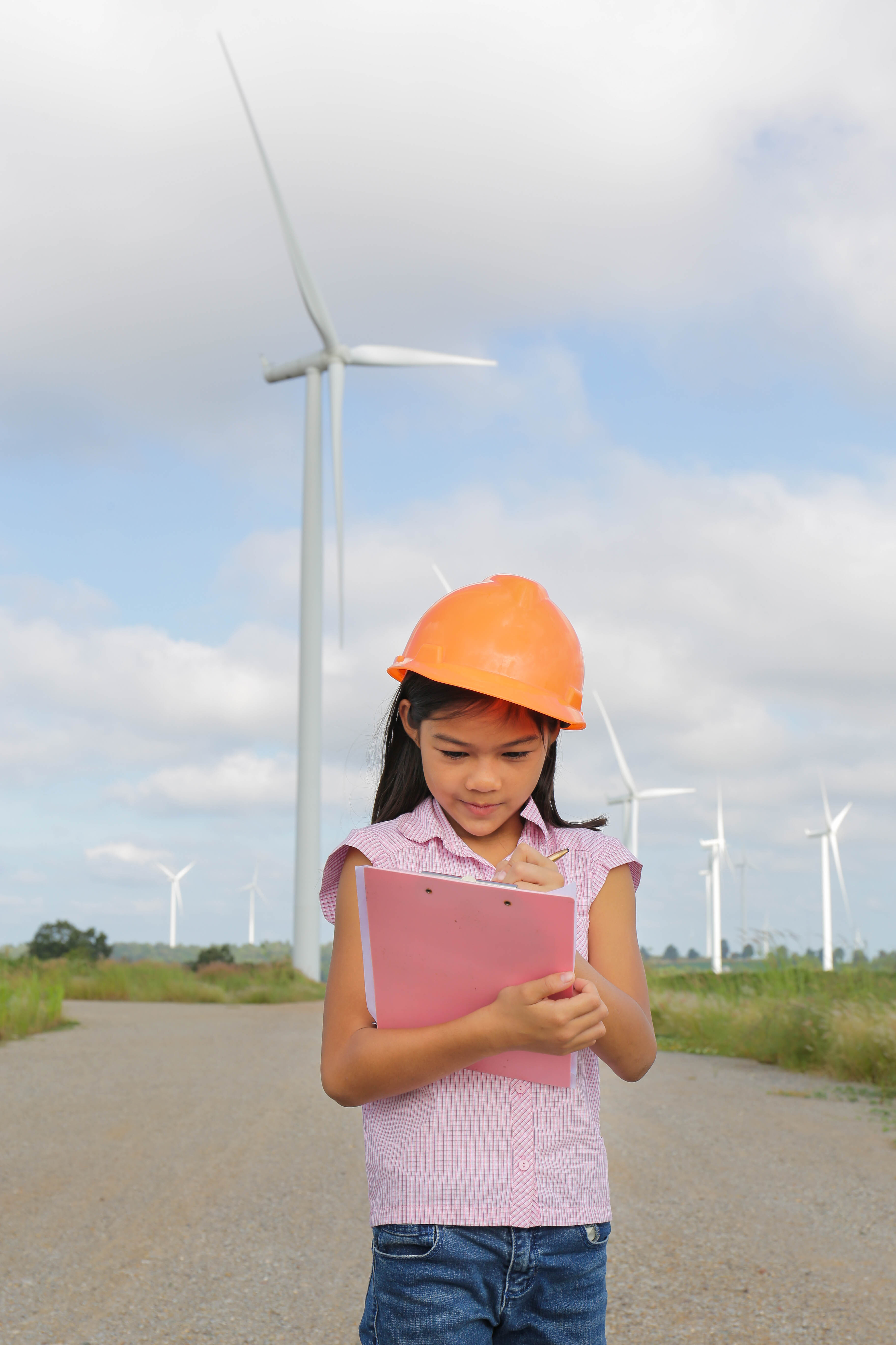 child standing in front of wind turbine wearing a hard hat and writing on a clipboard