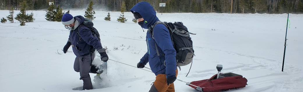 two people in snow gear, pulling sled with equipment through the snow