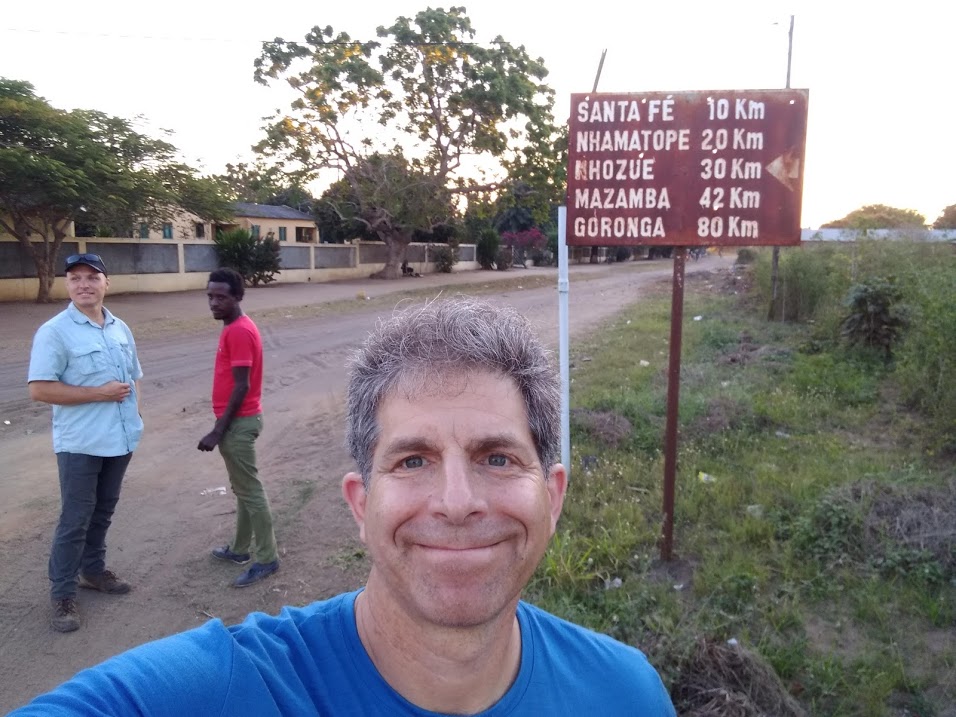 John Ziker taking a selfie in Mozambique, his traveling companions and a sign with distances to nearby cities in kilometers behind him.
