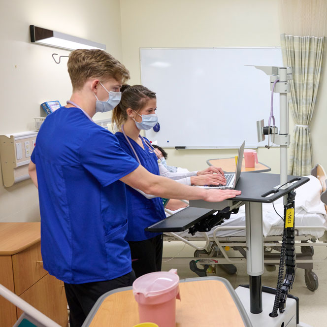 Two students in blue scrubs work a laptop computer next to a patient's bedside.