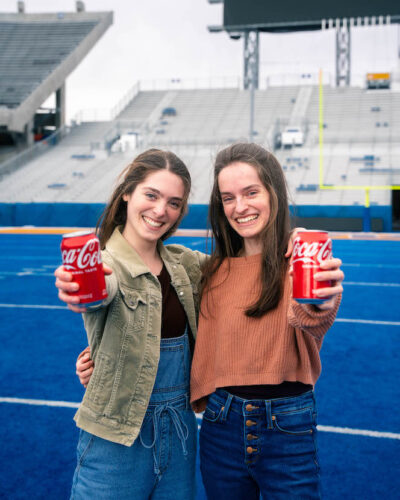 Women pose on the blue field of Boise State with cans of Coca-Cola