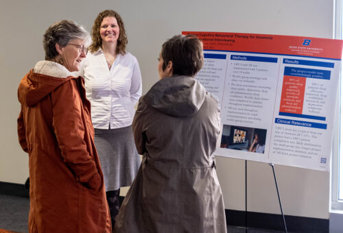 A doctoral student stands by her research poster and talks to two women about it.