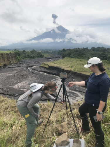 two young women stand outside in sight of an active volcano and set up camera on tripoc