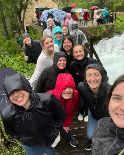 Students in raincoats stand in line on a bridge by a waterfall and smile.