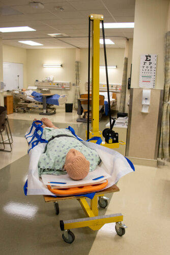 A manikin in a hospital gown lies on a crank-to-lift contraption in the Simulation Center.
