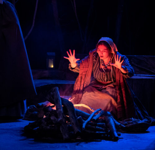 woman in Puritan costume kneels by a fire with hands up in the air, gesturing