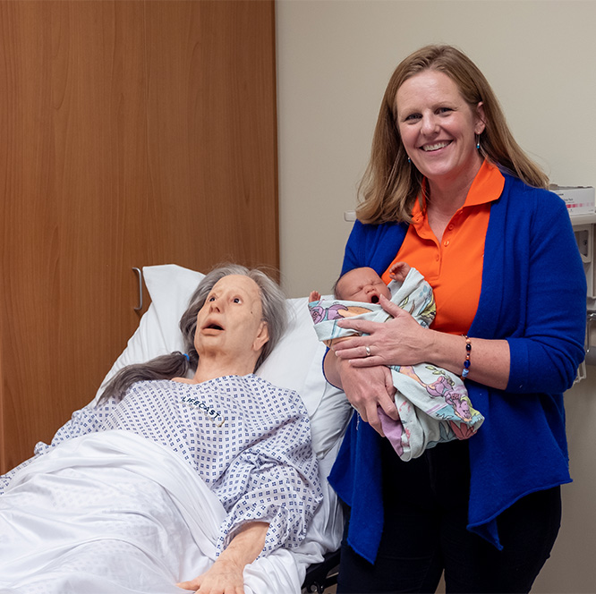 Kelley Connor smiles, holding a baby manikin next to an elderly manikin in bed.