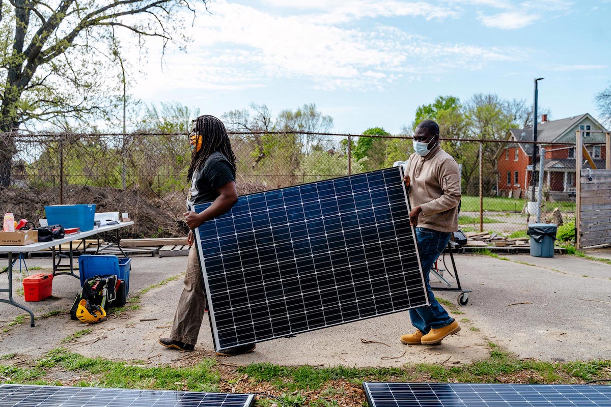 Two persons carry a solar panel in a residential neighborhood