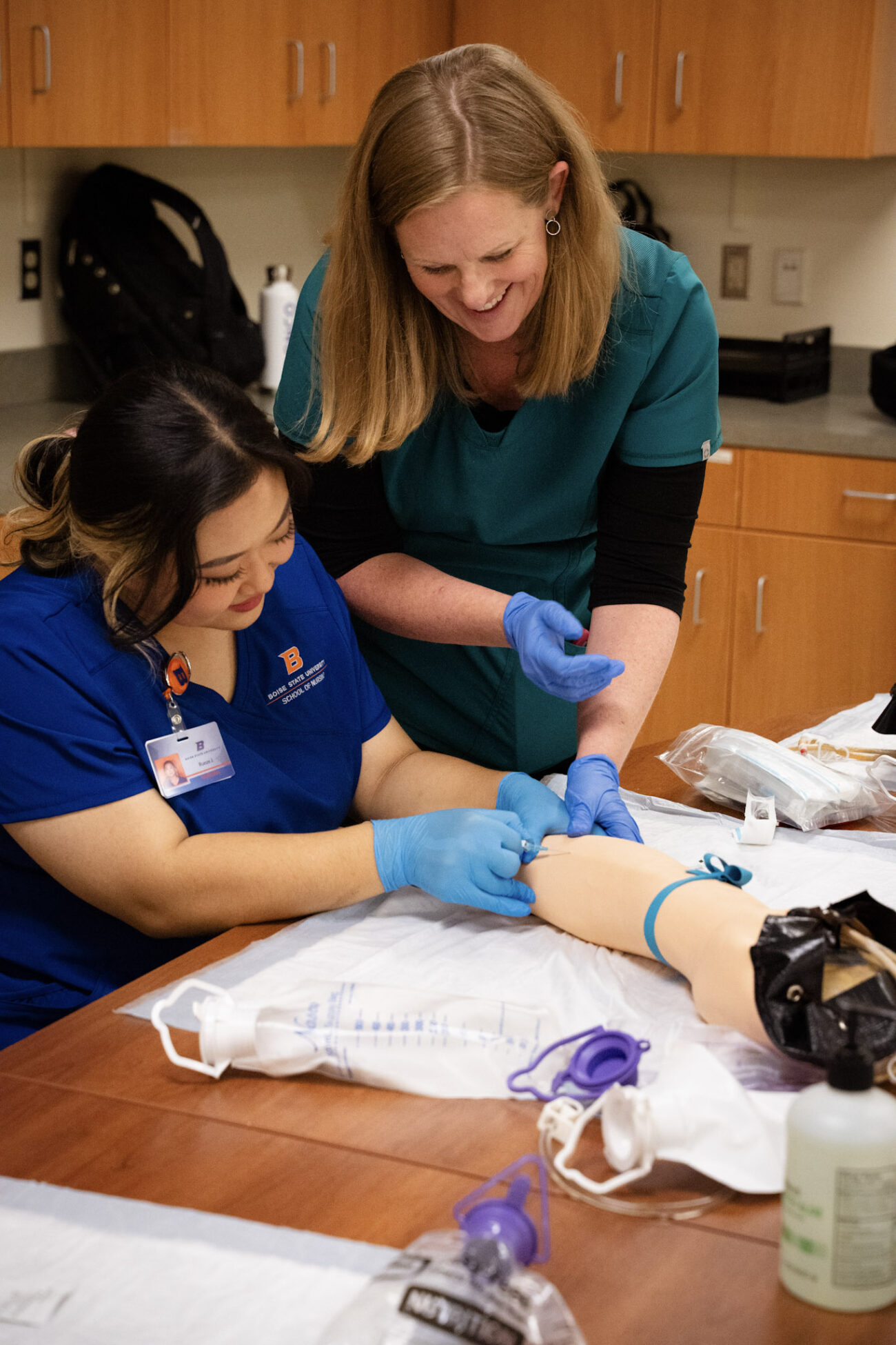 A nursing professor helps a student nurse insert an IV line in a manikin arm.