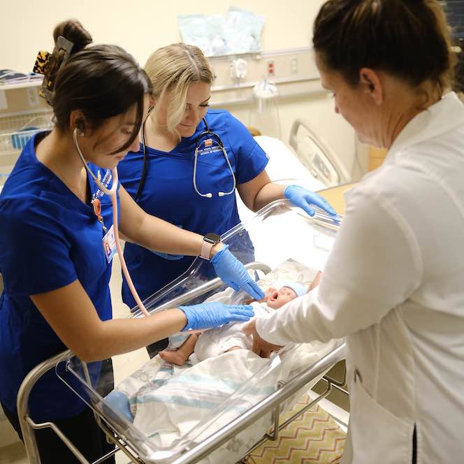 Two student nurses care for a baby manikin with a nurse in a white coat.
