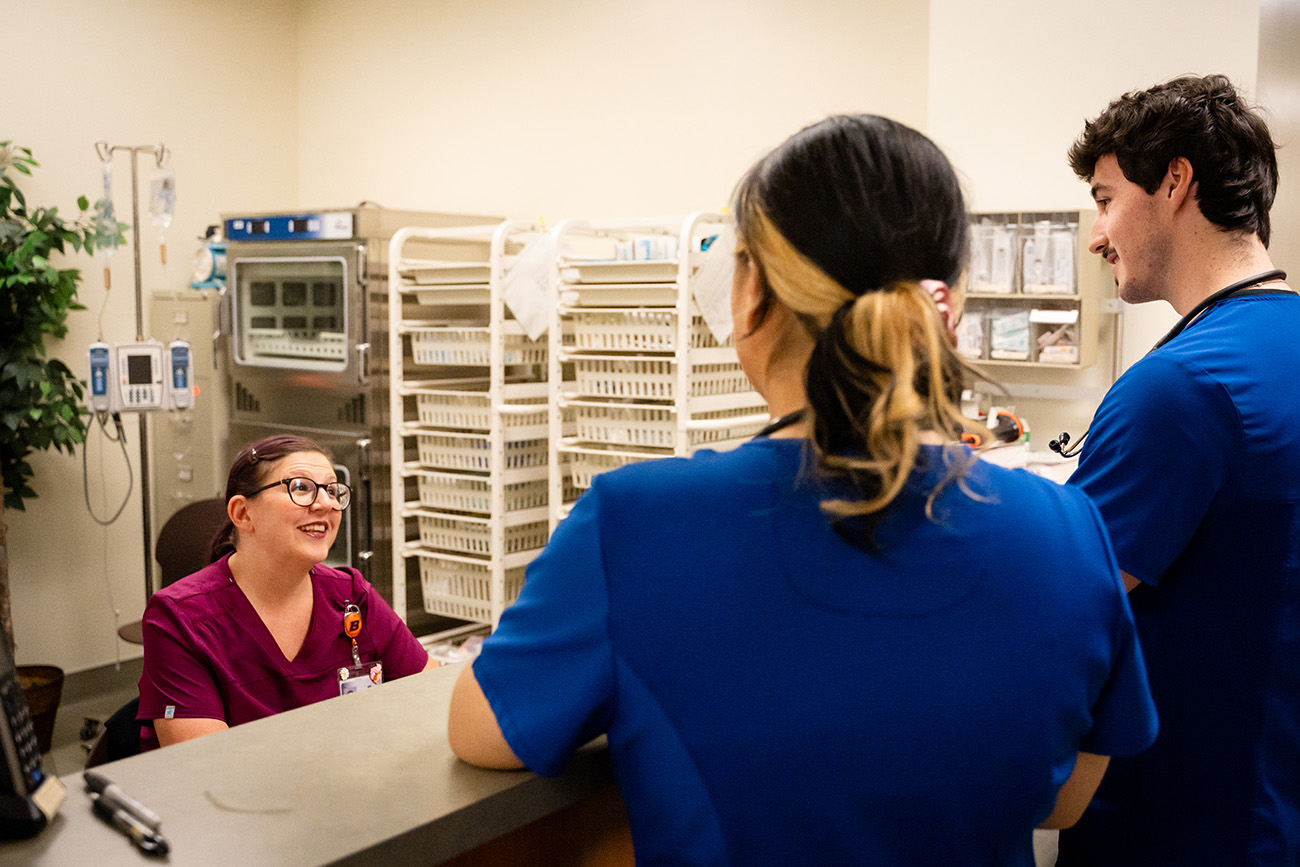 A smiling nurse sits at the nurses station desk looking up at two student nurses in scrubs.