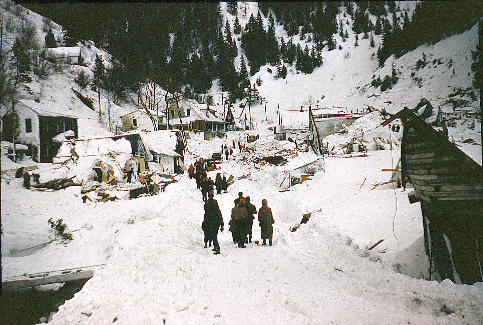 people walking up a plowed road to houses damaged by an avalanche