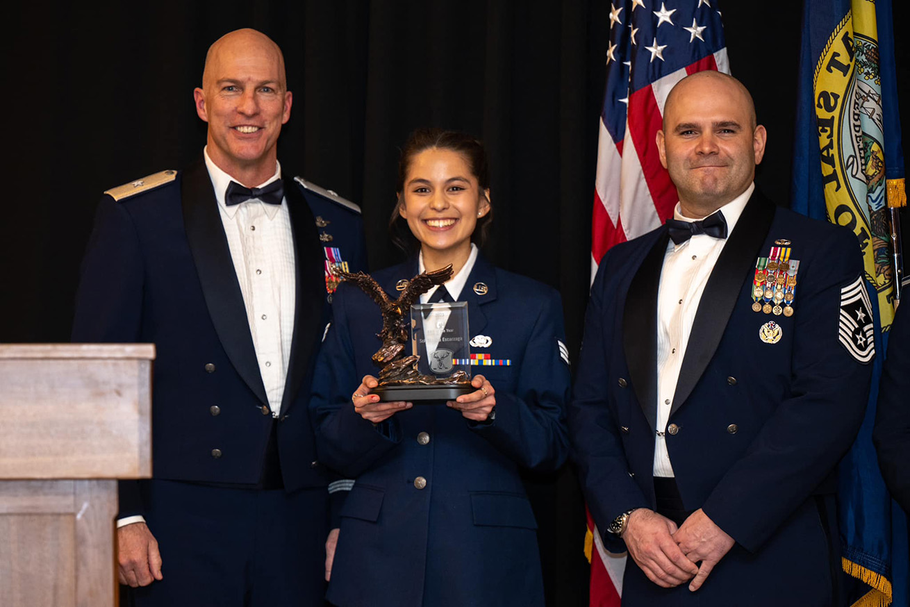 Escarzaga stands smiling between two senior Air National Guard officers and holds a large plaque.