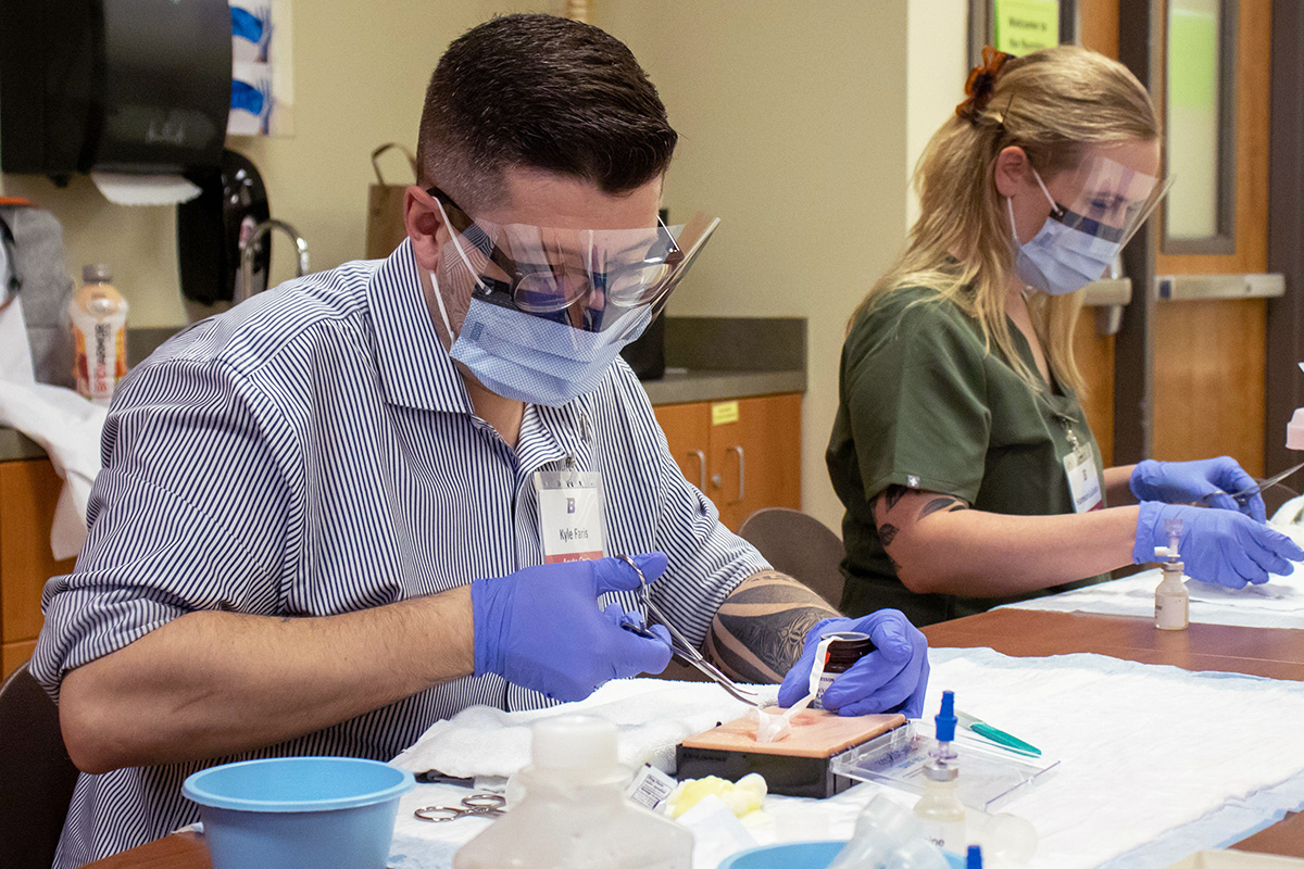 Two nurse practitioner students work at a table to practice simulated incisions in a sterile lab set up.