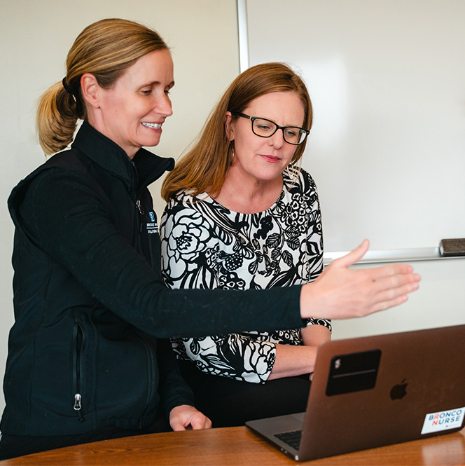 Kelley Connor looks on to Jenny Alderden's laptop as Jenny motions with her hand to explain what's on the screen.