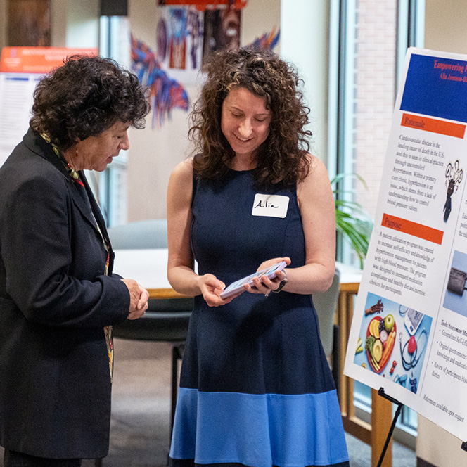 A doctoral nursing student stands by her research poster and demonstrates something on her phone to another woman.