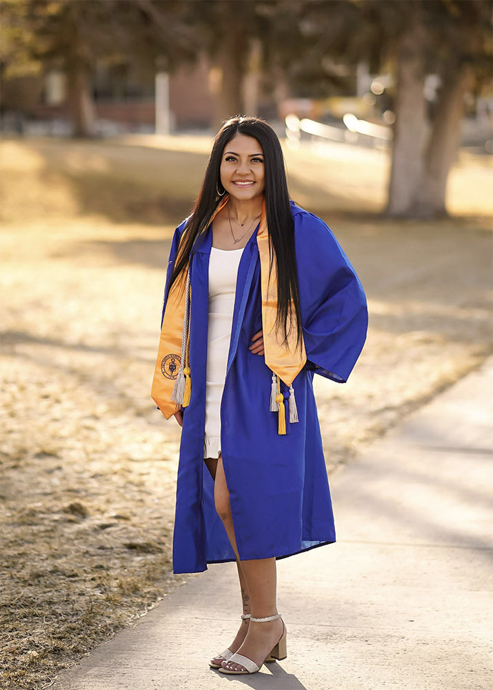 Alissa Godinez wears her regalia on a sunny sidewalk outside with trees in the background.