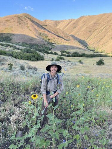 man stands wearing sunhat in Idaho rangelands, holding auger rod. Mountains in distance