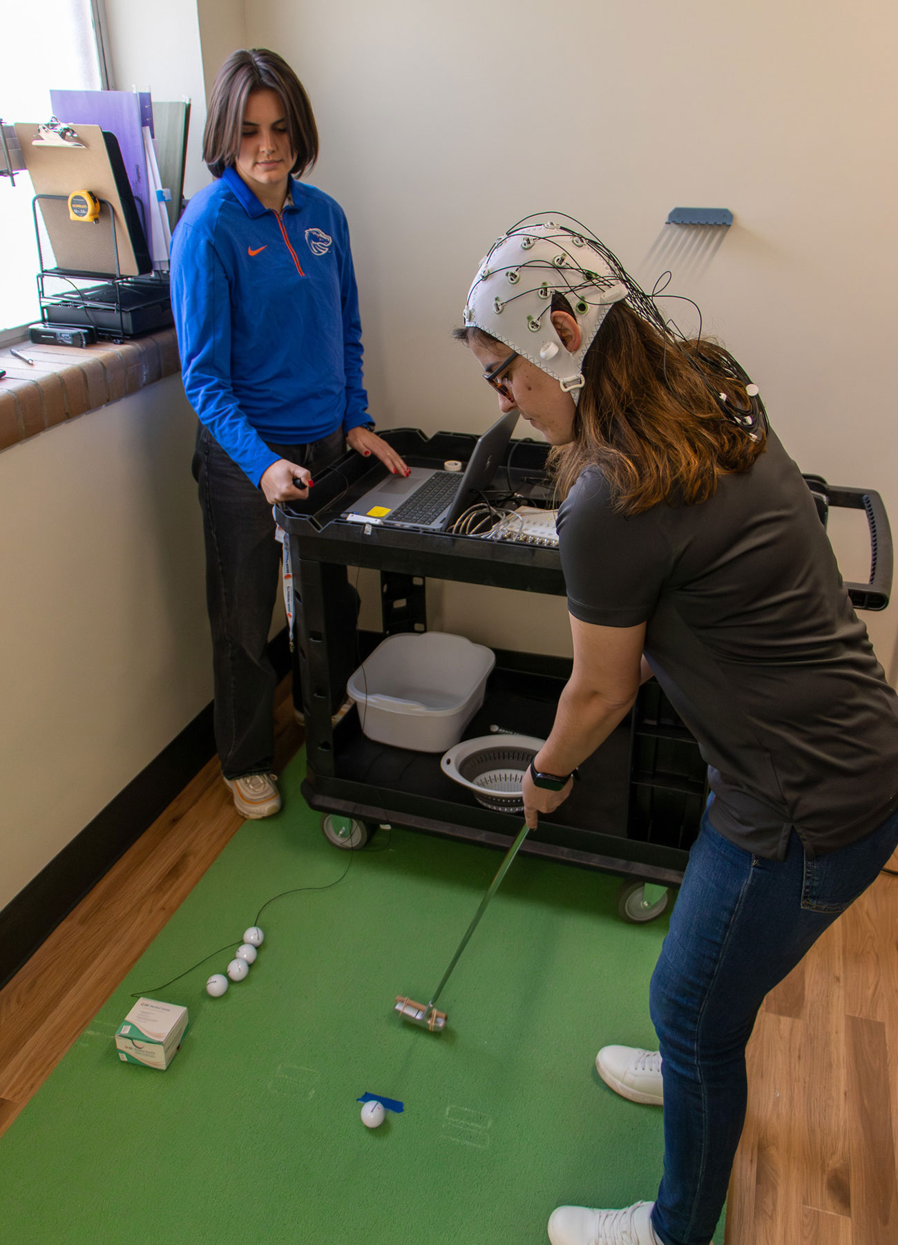 Kinesiology faculty Mariane Bacelar putts a golf ball while wearing an EEG hat, master's student Jet Taylor records the data on a laptop