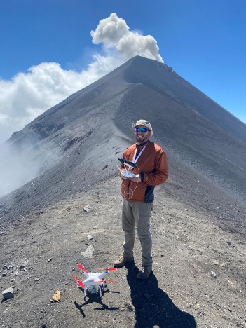 man stands on side of active volcano, holds remote control system for drone. Drone is at his feet