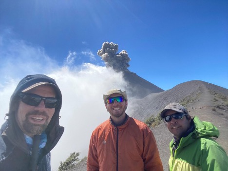 three men group together, behind them the volcano is actively spewing ash