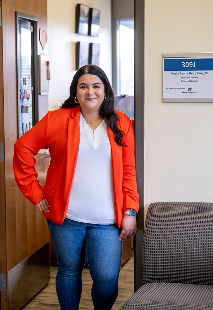 Maria Garcia De la Cruz leans on the doorframe by her office sign labeled with her name and "Academic Advisor".