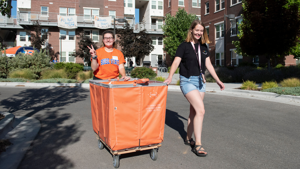 Two smiling students pulling a move-in carrier on wheels to their dorm rooms