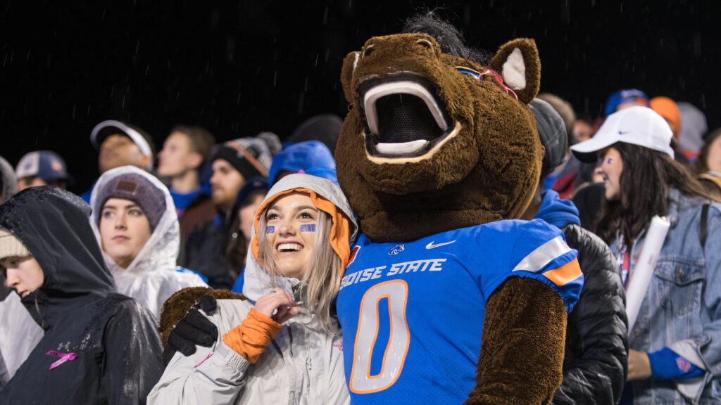 A student stands in the stadium seating with Buster Bronco mascot, embracing.
