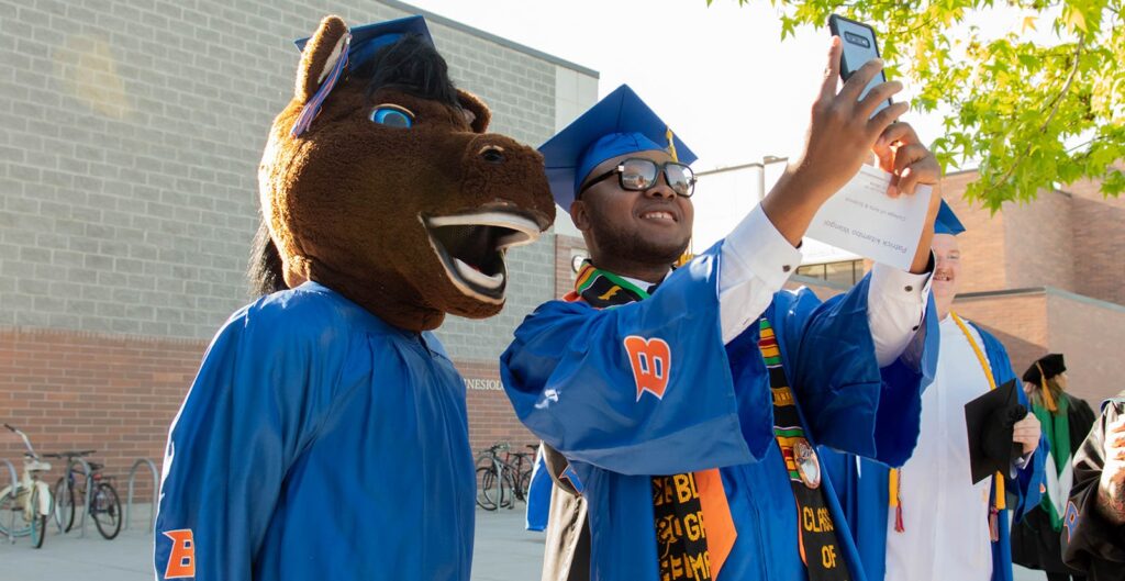 A graduating student taking a selfie with Buster Bronco.